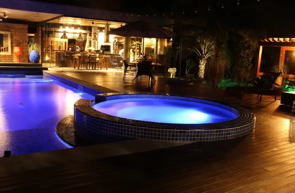 Piscina residencial iluminada à noite, com spa integrado e área de lazer externa ambientada com iluminação decorativa.