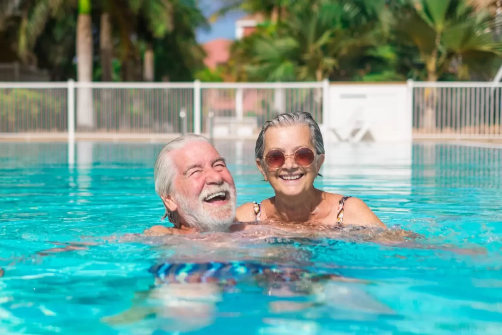 Casal de idosos sorrindo e se divertindo em uma piscina ao ar livre, em um ambiente seguro e acessível.
