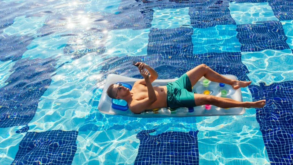 Homem deitado em uma boia dentro da piscina, mexendo em seu celular.