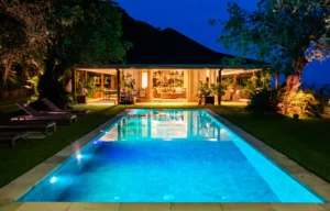 Piscina iluminada à noite em uma casa moderna com jardim e área externa aconchegante, refletindo luzes azuis na água cristalina.
