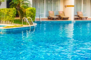 Piscina externa com água azul cristalina, escada de aço inox, espreguiçadeiras e paisagismo em um ambiente residencial ou hoteleiro moderno.